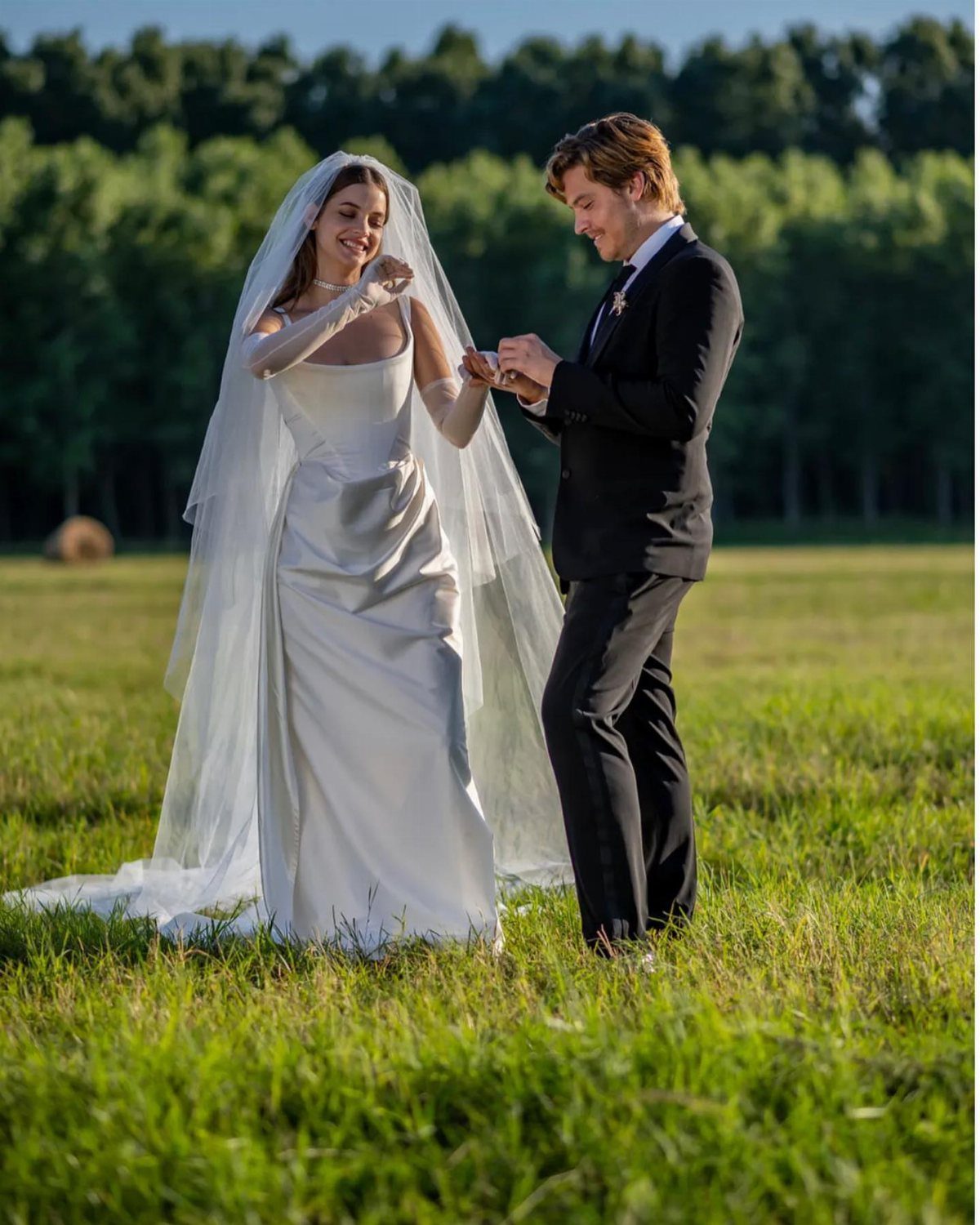 Bride in white satin dress and groom in black tuxedo in a field