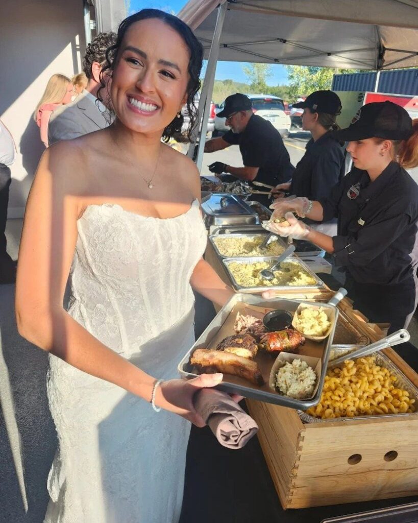 Bride in white lace dress holding a tray of barbecue ribs and sides.