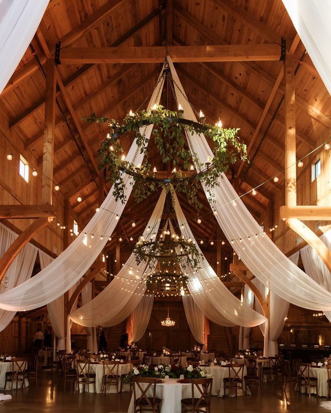 Wooden barn interior decorated with white fabric drapes, greenery wreaths, and wedding tables.