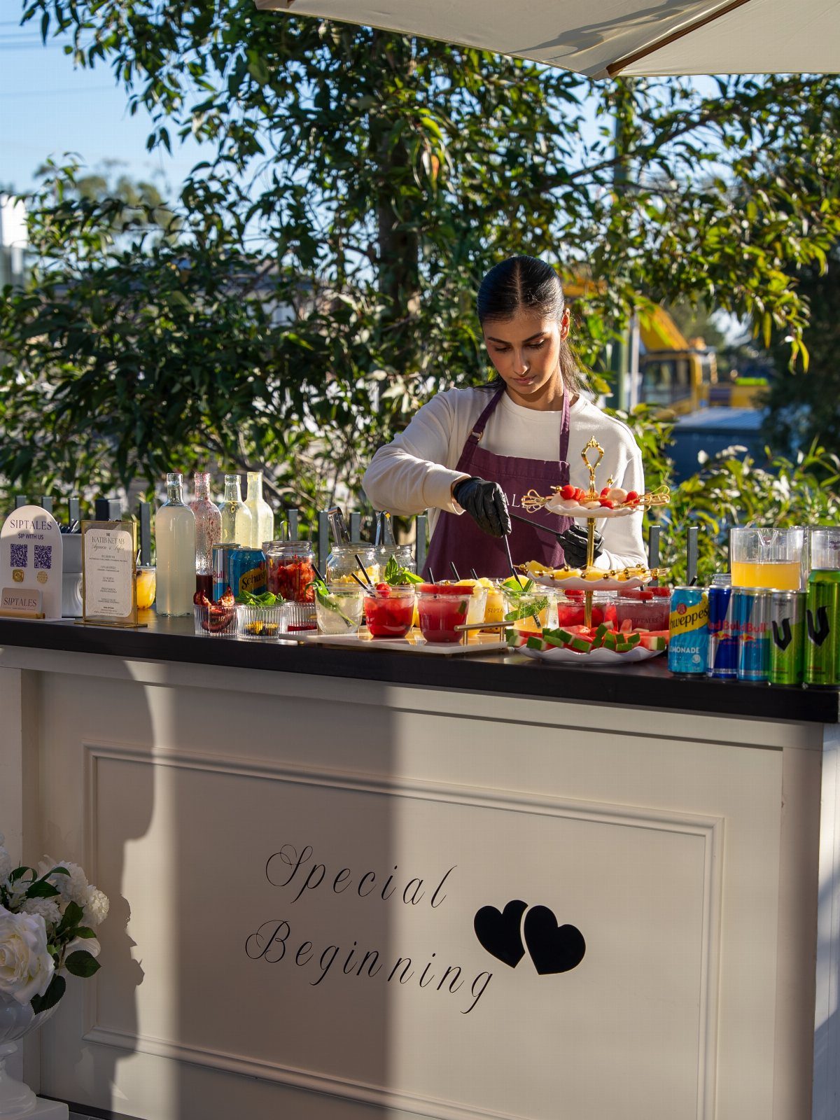 Bartender preparing colorful cocktails at a white wedding bar outdoors under a white umbrella