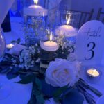 Floating white candles in glass cylinders with white roses and greenery on wedding table.