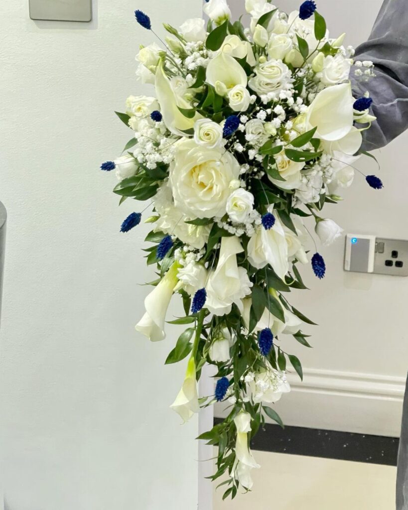 Cascading bridal bouquet with white roses, calla lilies, and small blue accent flowers.