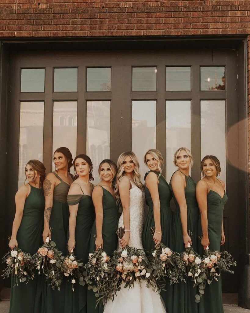 Bride in white gown with bridesmaids in dark green dresses holding greenery bouquets.