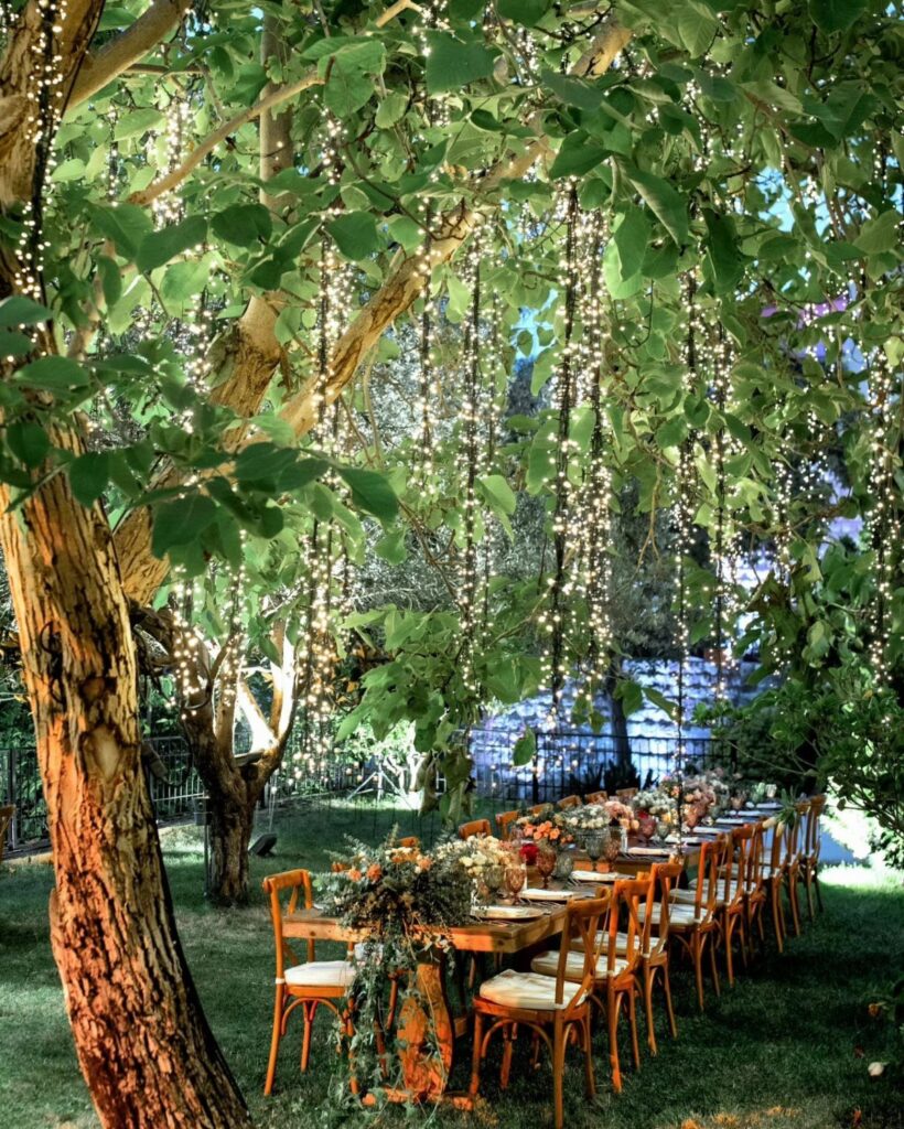 Wooden dining table under trees draped with hanging strings of warm fairy lights.