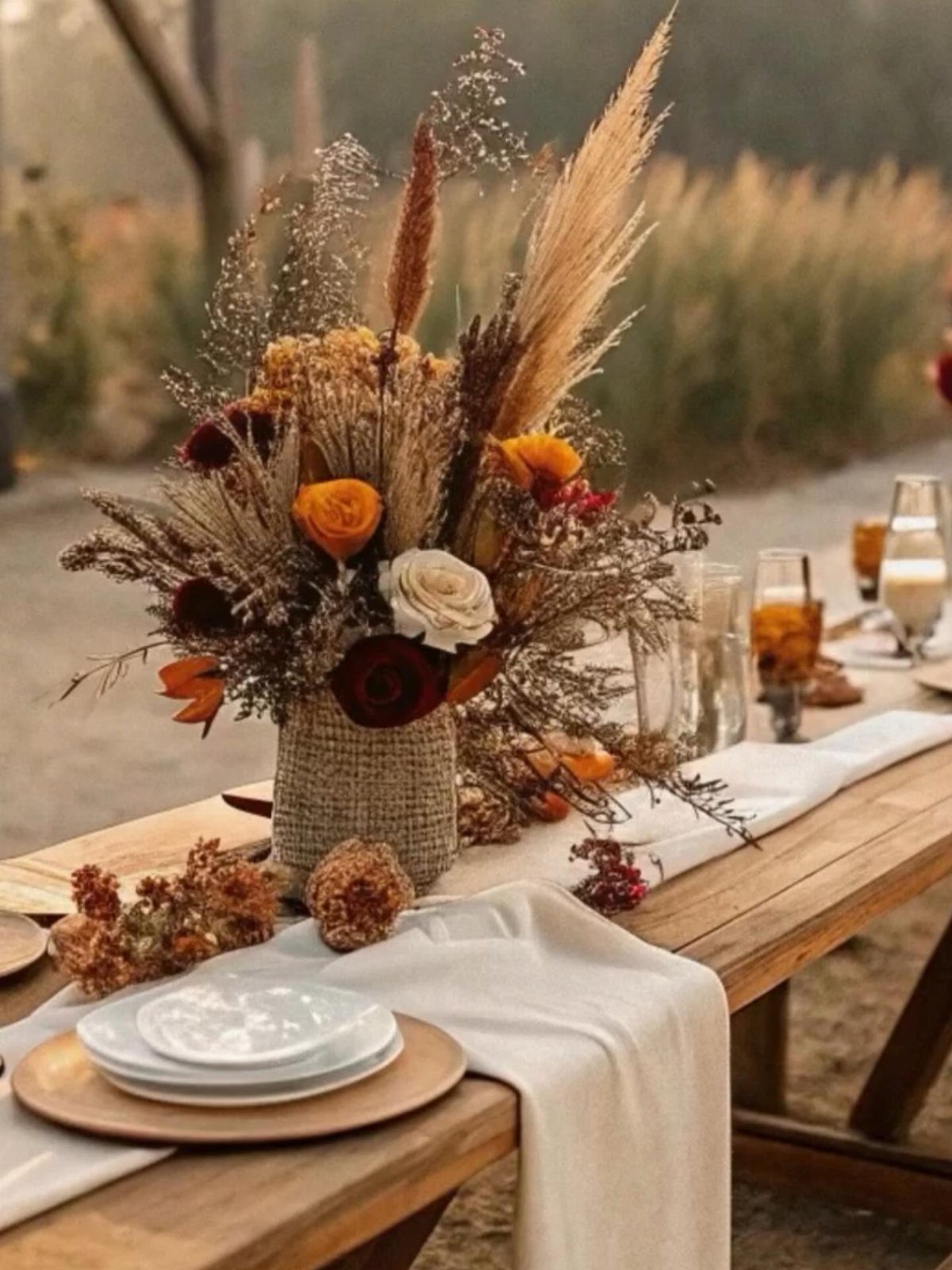 Autumnal floral centerpiece with orange roses and pampas grass on a wooden wedding table.