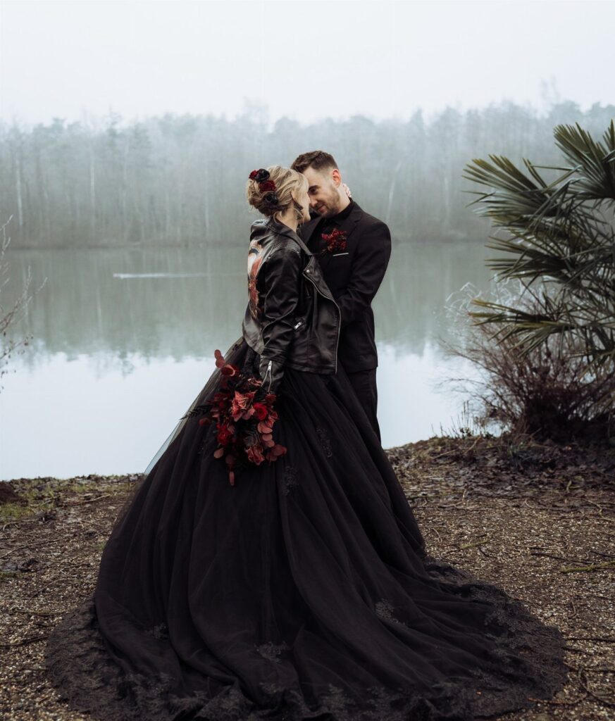 Couple in gothic black wedding attire posing by a misty lake with red flowers