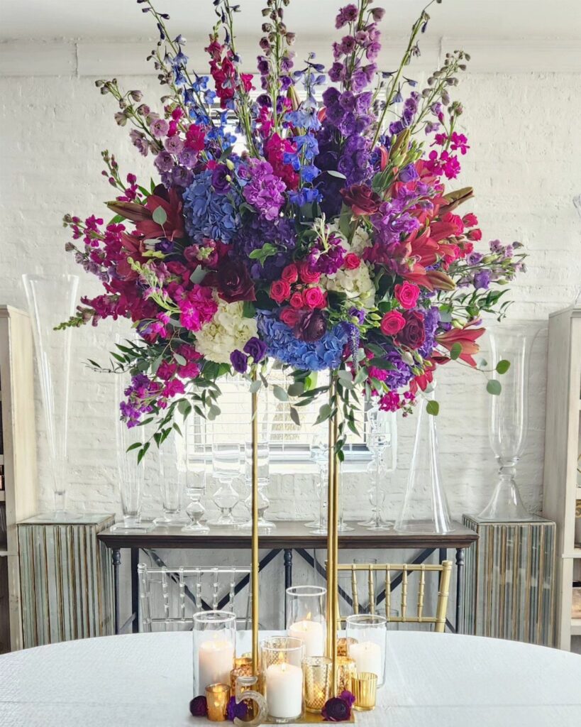 Tall gold floral centerpiece with jewel-toned purple and blue flowers on white table