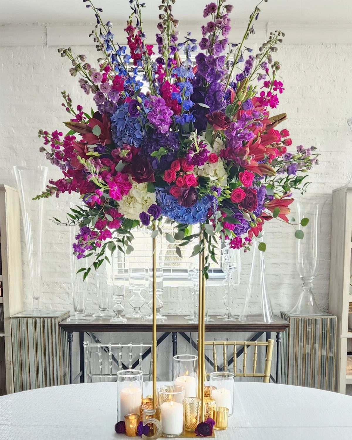 Tall gold floral centerpiece with jewel-toned purple and blue flowers on white table