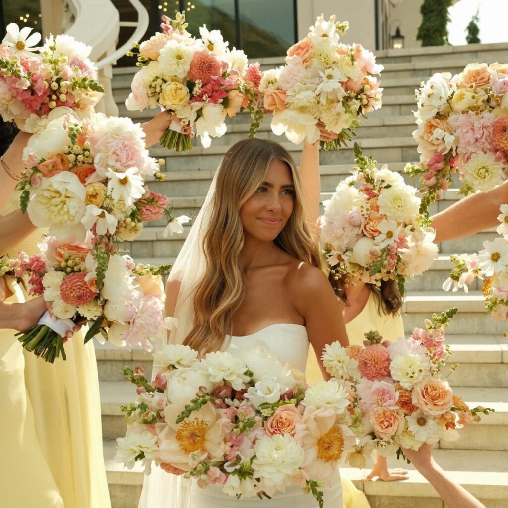 Bride in white gown surrounded by bridesmaids with yellow dresses and peach floral bouquets.