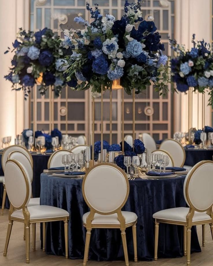 Navy blue velvet table setting with gold chairs and tall blue floral centerpieces.