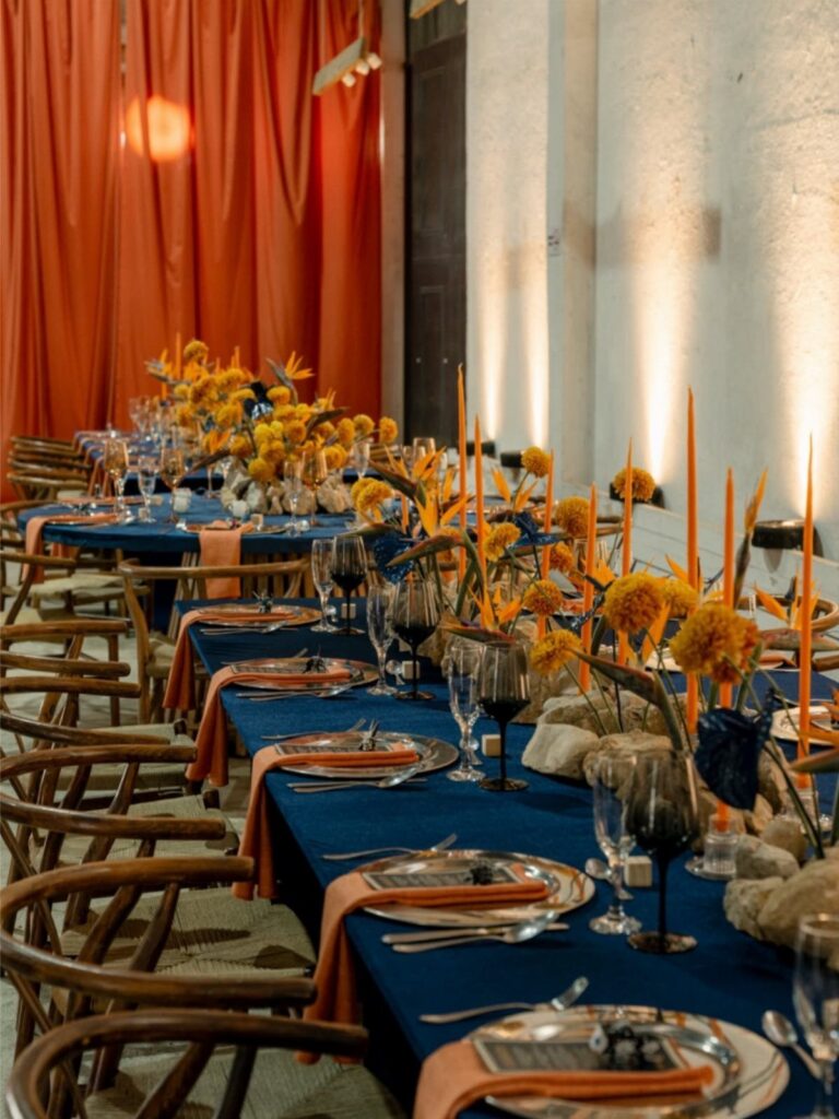 Dining table with blue tablecloth, orange napkins, and orange flowers for a wedding.