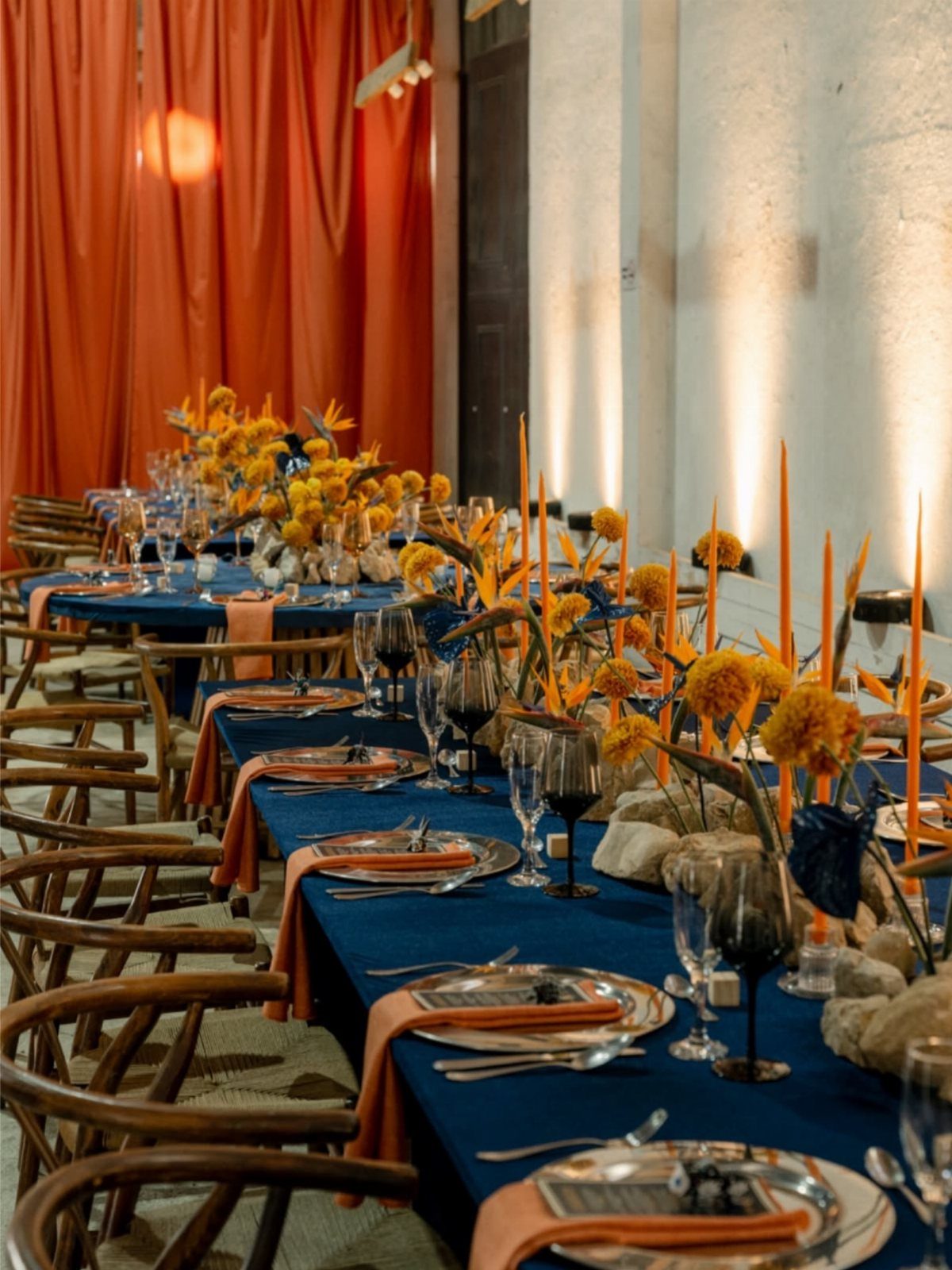 Dining table with blue tablecloth, orange napkins, and orange flowers for a wedding.