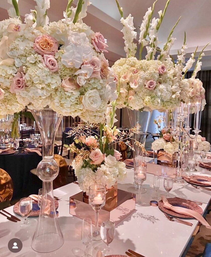 Rose gold wedding table setting with tall white and pink floral centerpieces and metallic accents.