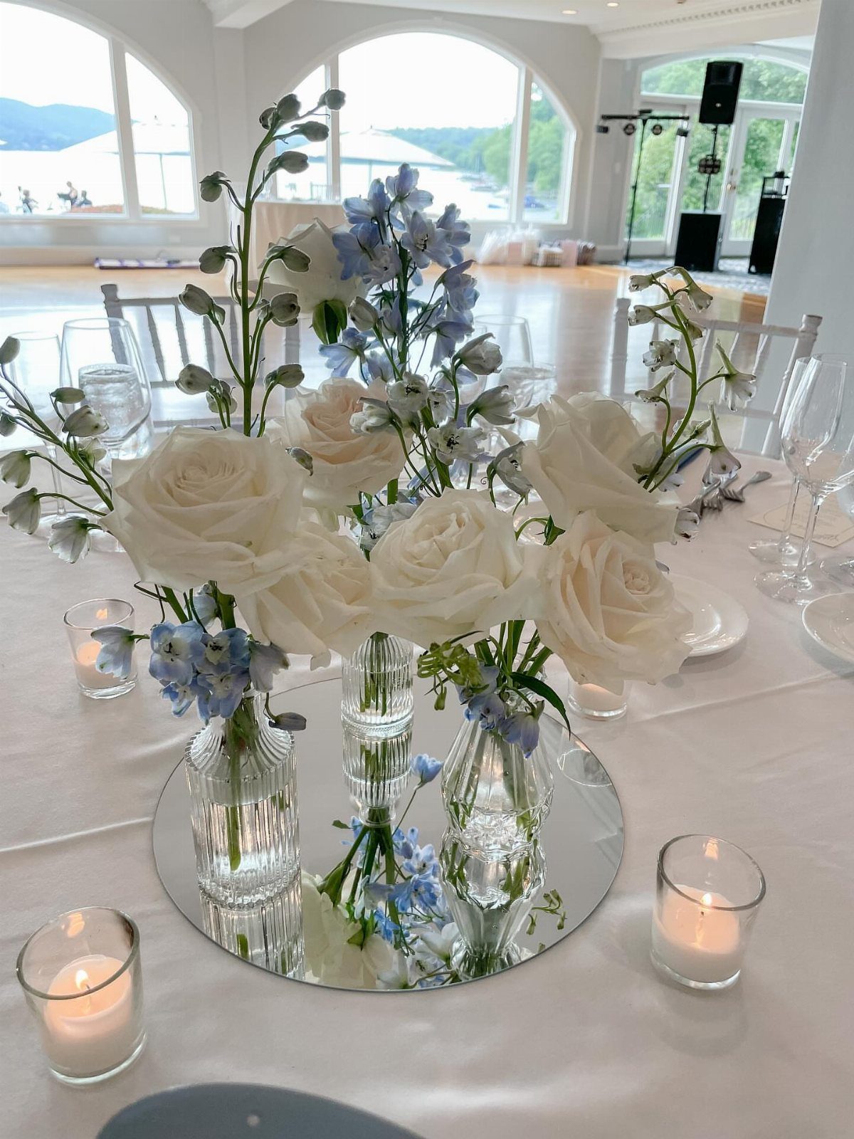 White roses and blue flowers in glass vases on a mirrored table centerpiece.