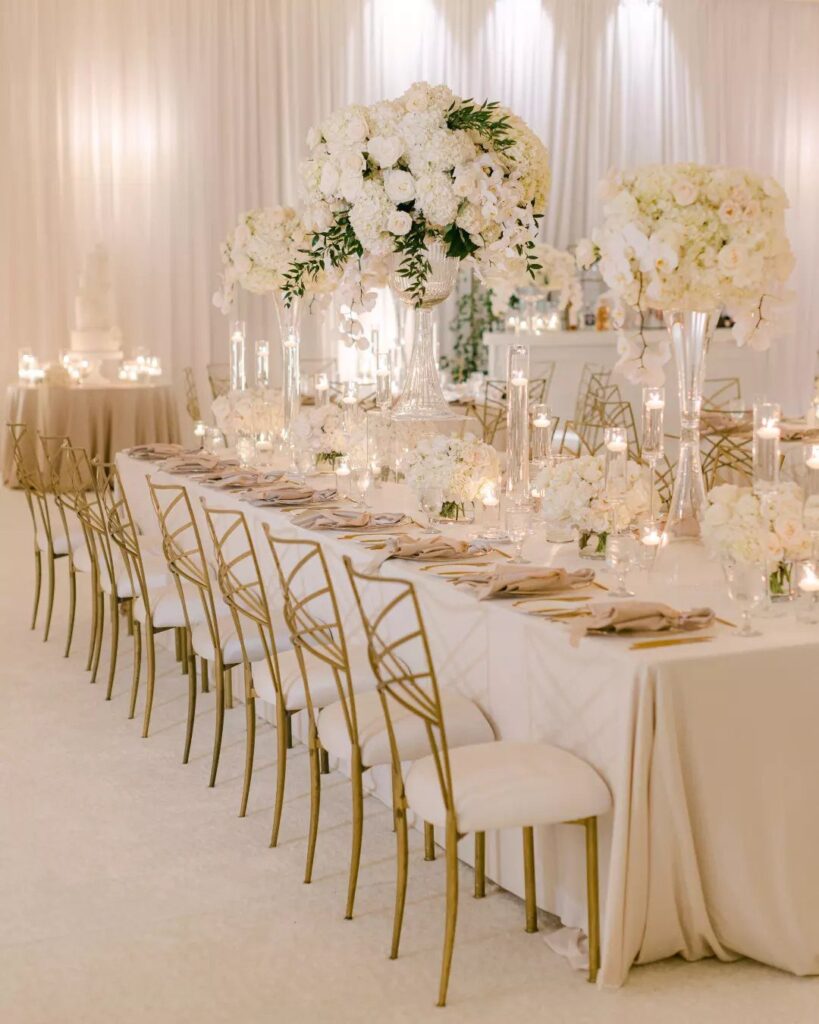 Ivory banquet table with gold chairs and white floral centerpieces in a ballroom.