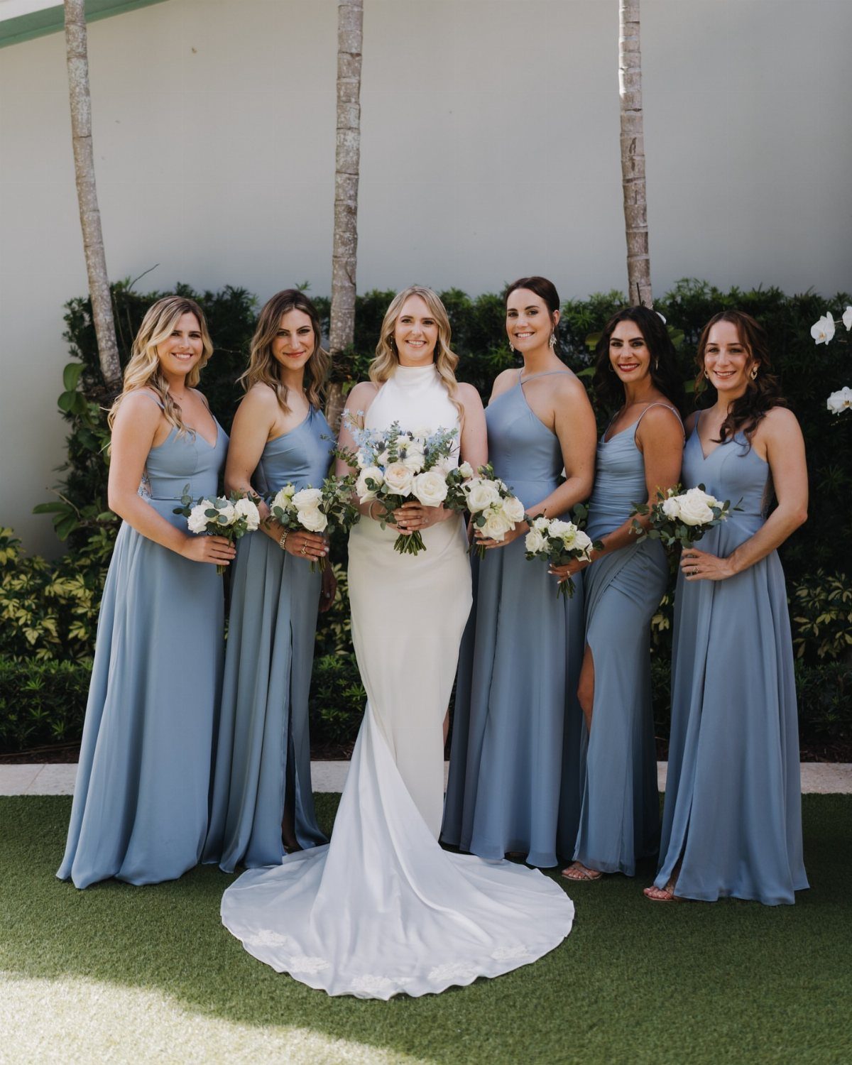 Bride in white gown surrounded by bridesmaids wearing dusty blue dresses with white bouquets.