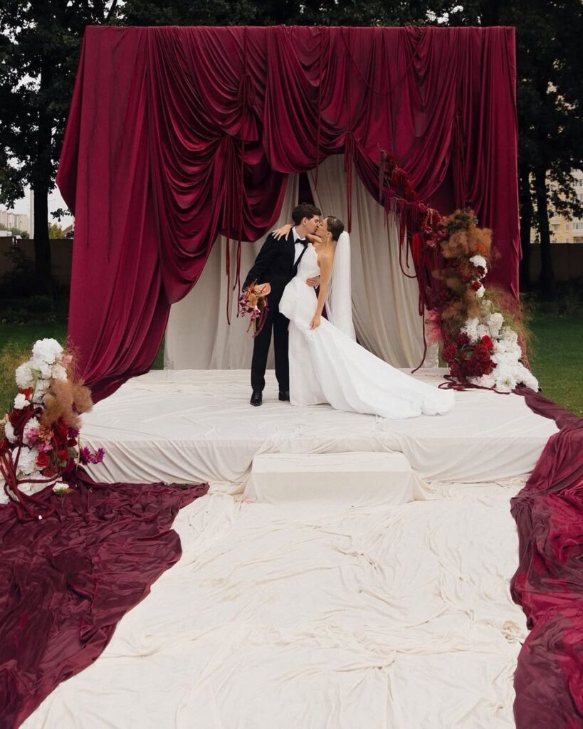 Bride and groom kissing before a large maroon fabric backdrop and floral arrangements.