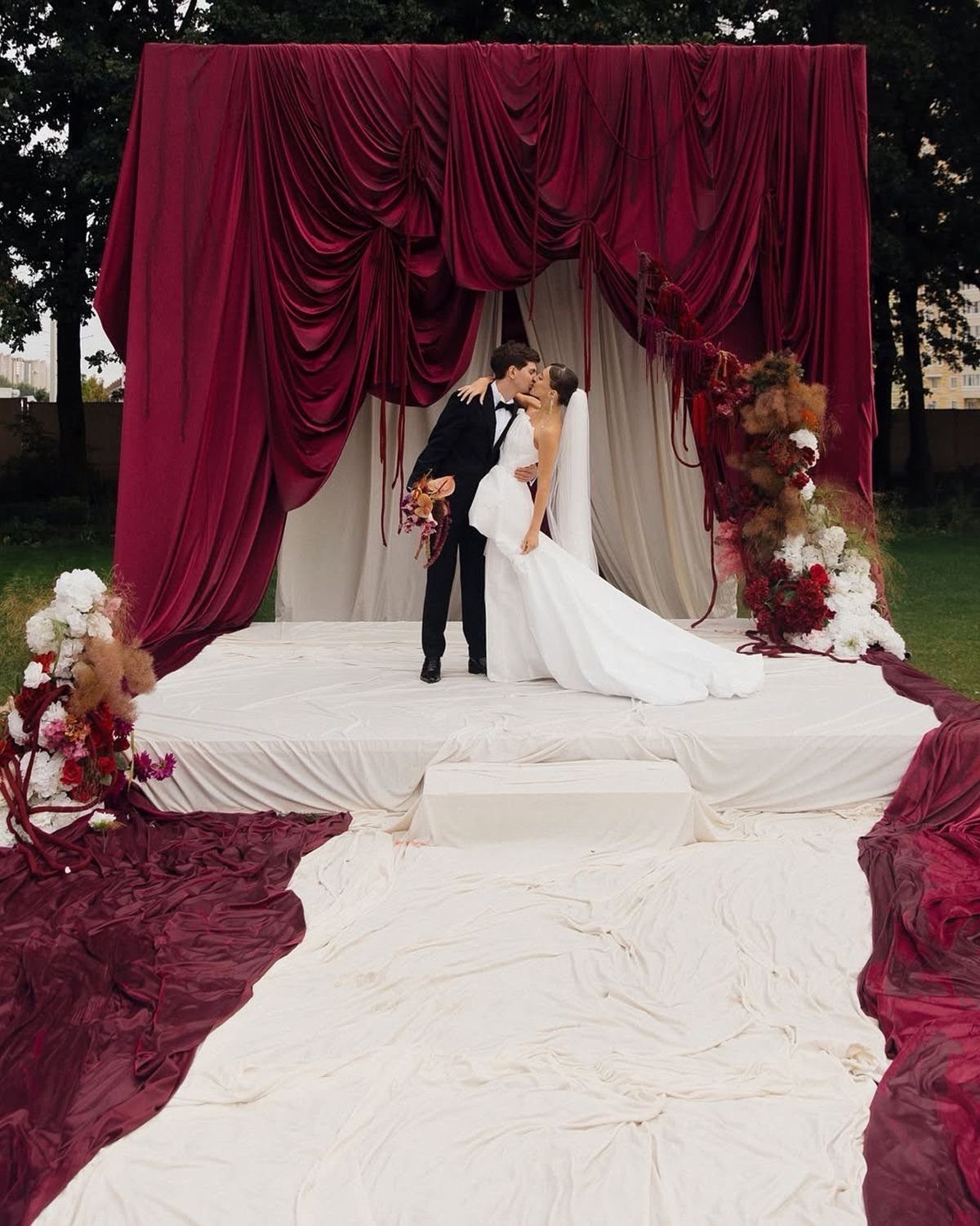 Bride and groom kissing before a large maroon fabric backdrop and floral arrangements.
