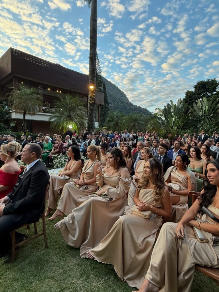 Bridesmaids in champagne satin dresses sitting outdoors at a wedding ceremony.