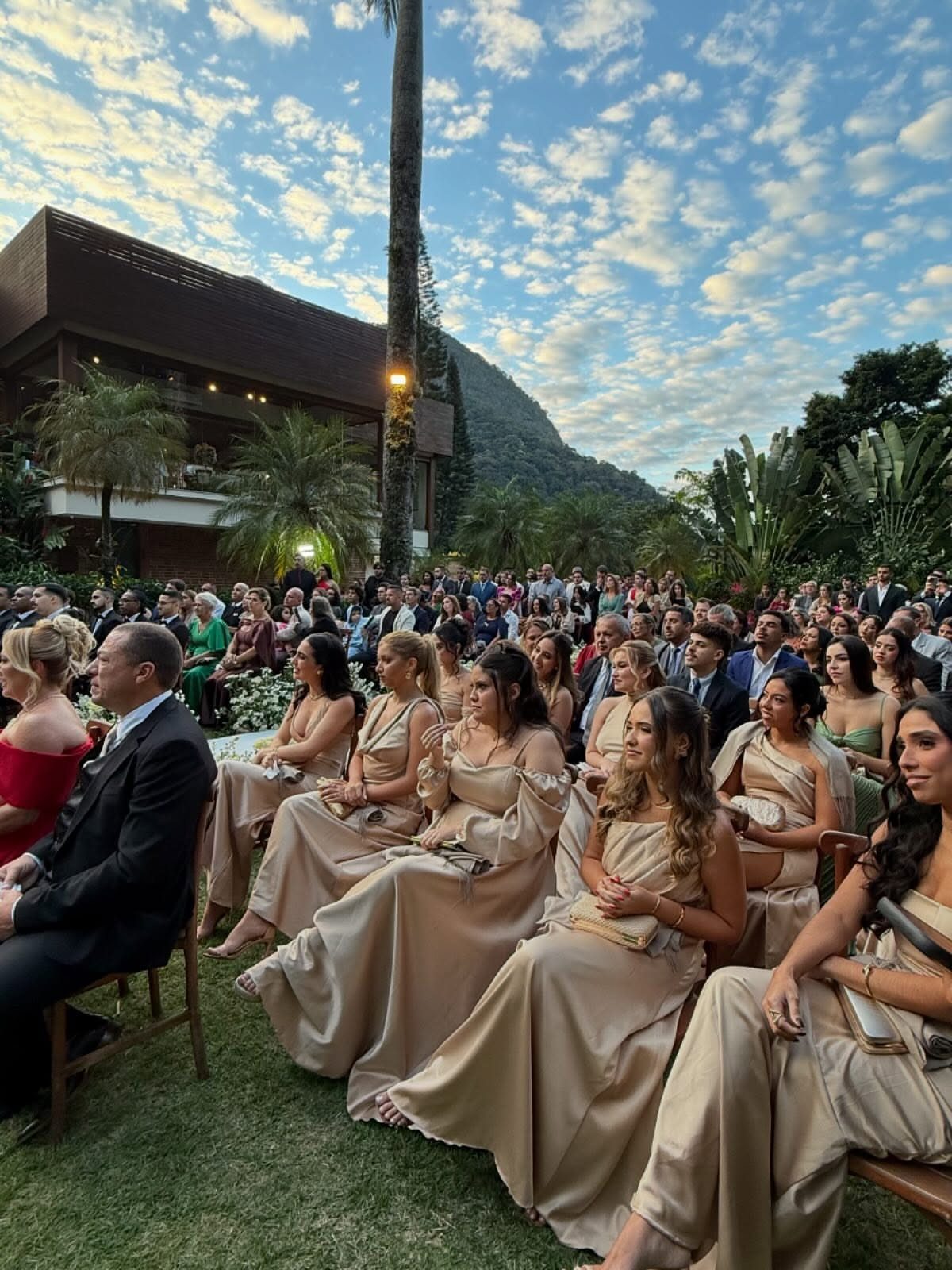 Bridesmaids in champagne satin dresses sitting outdoors at a wedding ceremony.