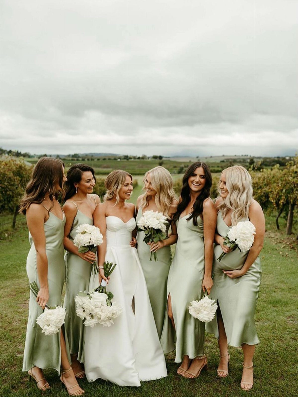 Bride in white gown with bridesmaids in sage green dresses holding white bouquets.
