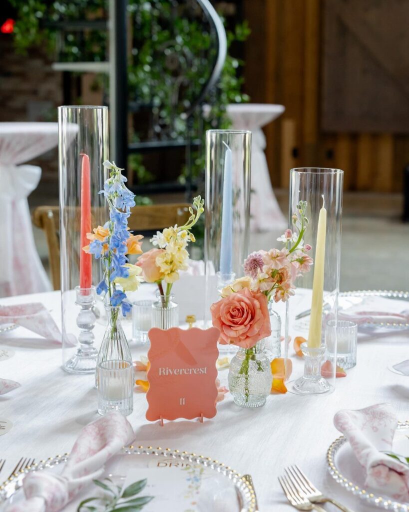 Colorful taper candles in glass cylinders with floral bud vases on white table.