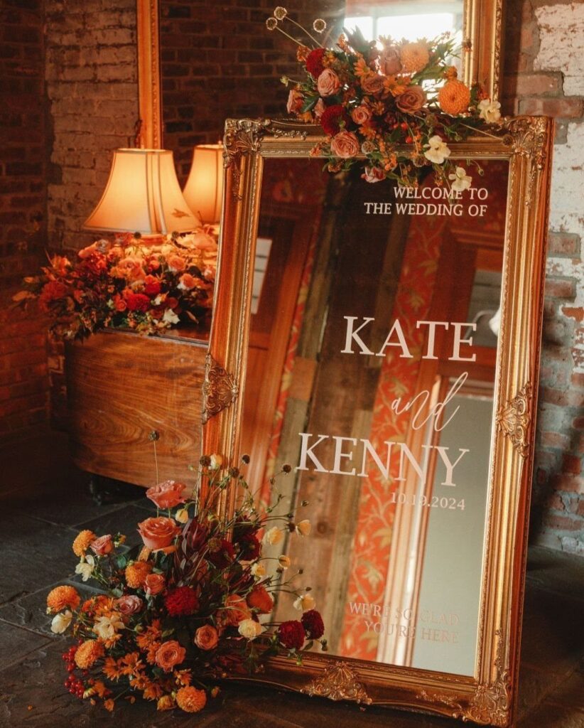 Gold ornate mirror wedding welcome sign decorated with autumn orange and red flowers.