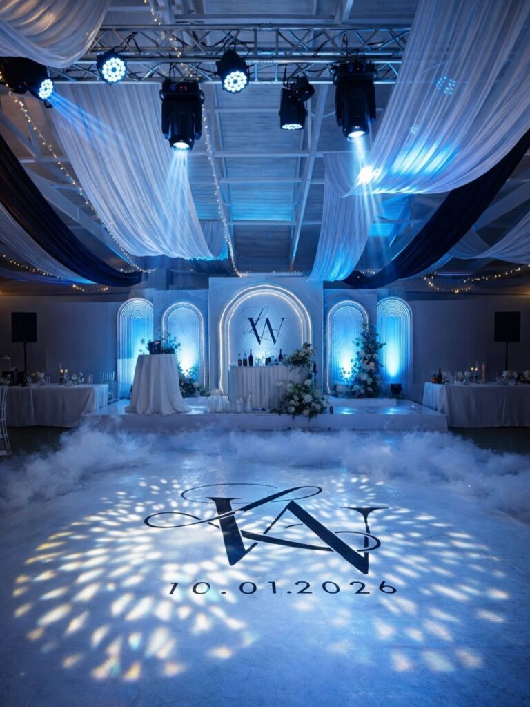Wedding reception dance floor with blue lighting, white ceiling drapes, and a projected monogram.