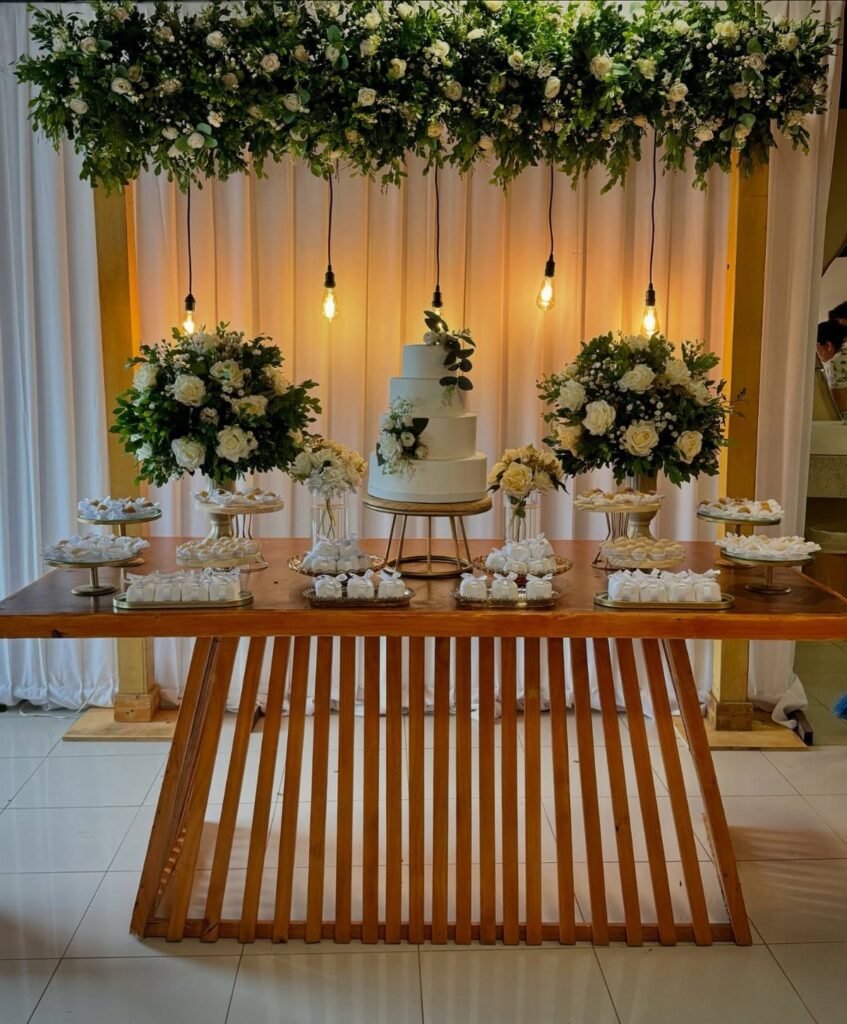 Wooden wedding dessert table with white cake, floral arrangements, and hanging greenery installation.