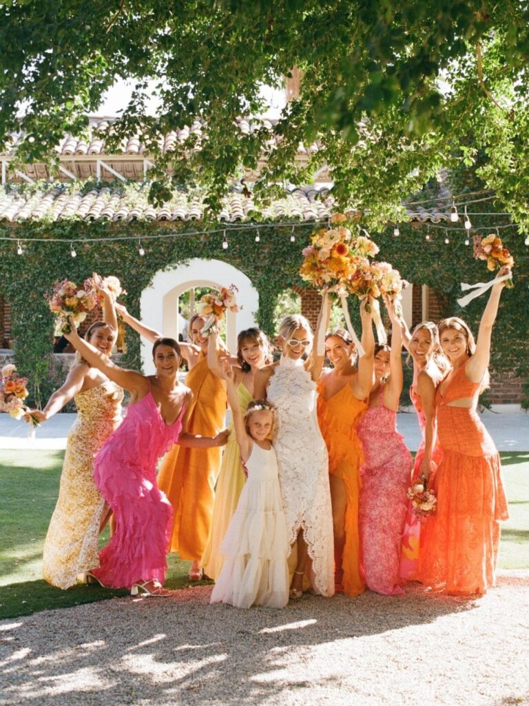 Bride and bridesmaids in colorful dresses cheering outdoors with floral bouquets.