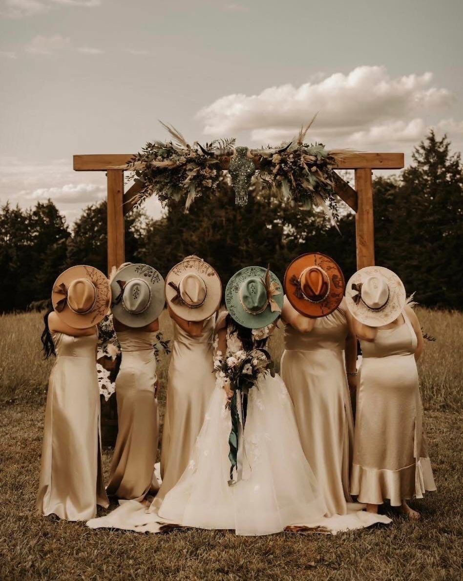 Bride and bridesmaids in champagne dresses holding western hats before a wooden arch.