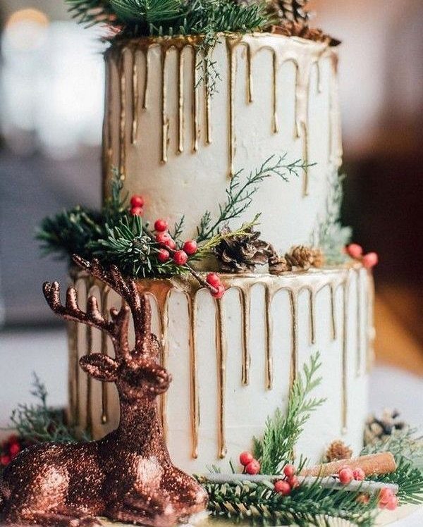 Two-tier white cake with gold drips, evergreen sprigs, red berries, and a glittery reindeer.